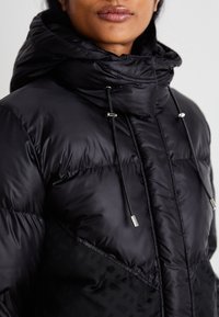 Mustat puffatakki, jossa on huppu, kiristysnauhat, sileä pinta ja kontrastivärinen kuvioitu paneeli alareunassa.