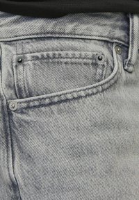 Primer plano de un bolsillo de jeans de denim gris claro con remaches negros visibles y detalles de costura.