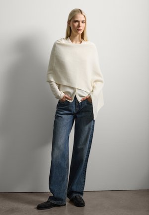 Poncho texturizado en color crema, colocado sobre una blusa blanca, combinado con jeans holgados de color azul claro y zapatos negros. Diseño sencillo y minimalista.