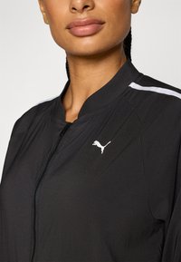 Black zip-up jacket with a high collar, featuring white stripes on the shoulders and a small white logo on the chest. Smooth texture.
