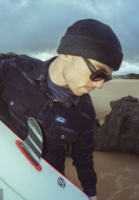 Chemise à carreaux noirs avec un patch, bonnet noir, lunettes de soleil, et une planche de surf avec un bord rouge et une nageoire rayée, sur une plage de sable avec des rochers.