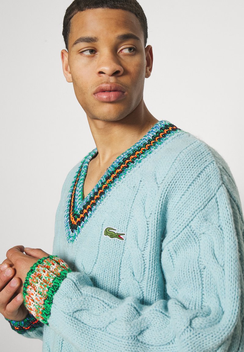 Giovane uomo che guarda di lato, indossa un maglione a maglia leggera di colore azzurro con dettagli colorati e un logo di coccodrillo sul petto.