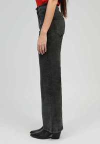 Femme portant un jean noir taille haute, des bottes à talons pointues noires et un haut rouge, se tenant de côté devant un fond en clair.