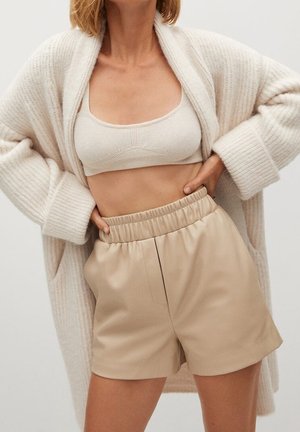 Femme portant un bralette en maille beige, un cardigan crème oversize et un short en cuir beige taille haute avec une ceinture élastique. Mains sur les hanches.