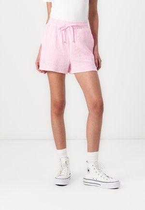 Person iført lyserøde teksturerede shorts med snøre, hvide høje sneakers og hvide sokker mod en ensartet hvid baggrund.