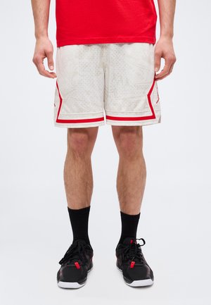 Homme portant un short de sport blanc avec des bandes rouges, des chaussettes noires et des baskets noires, debout devant un fond clair uni.