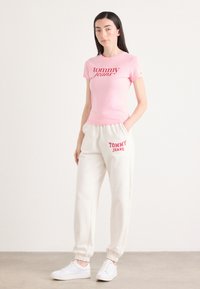 Roze katoenen T-shirt met "tommy jeans" logo, gecombineerd met crèmekleurige joggingsbroek met rode "TOMMY JEANS" letters, en witte sneakers.