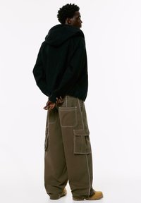 Uomo di profilo che indossa un hoodie nero, pantaloni cargo marroni oversize con cuciture bianche e stivali beige su sfondo bianco.