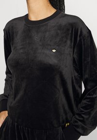 Žena v černé sametové mikině Adidas s jemnými embosovanými trojlístkovými vzory a malým zlatým logem Adidas na hrudi.