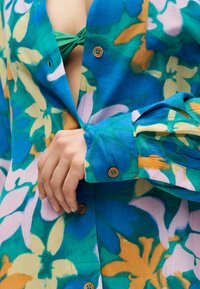 Χέρι ρυθμίζει το μανίκι ενός μπλε, πράσινου και πορτοκαλί πουκάμισου με λουλούδια και κουμπιά, με ένα πράσινο τοπ μπικίνι ορατό από κάτω.