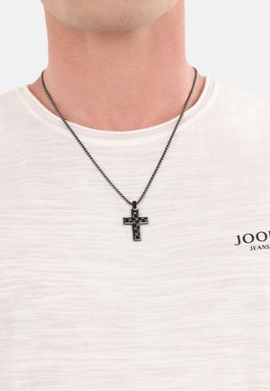 Black cross pendant necklace on a dark chain, worn over a light-colored shirt with a textured pattern. Cross features a geometric design.