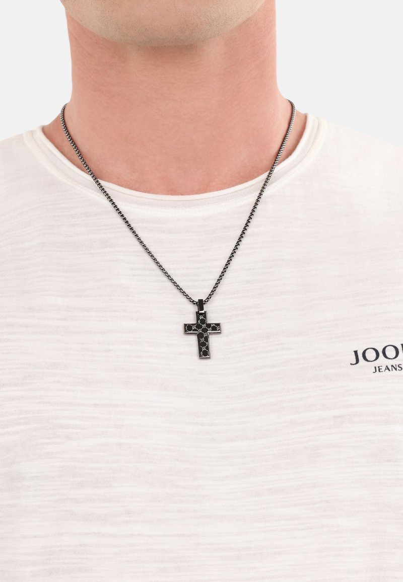 Zwarte kruisketting aan een donkere ketting, gedragen over een lichtgekleurde shirt met een gestructureerd patroon. Het kruis heeft een geometrisch ontwerp.