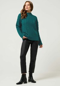 Türkisfarbener Kabelstrickpullover mit langen Ärmeln und hohem Kragen, kombiniert mit schwarzen Lederhosen und Blockabsatz-Stiefeletten.