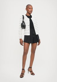 White knitted jacket with horizontal stripes, black top, black pleated shorts, and strappy black heels; holding a small black bag.