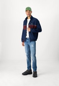 Rompevientos azul marino y granate con cremallera, bolsillos y puños elásticos sobre una camiseta blanca. Combinado con jeans desgastados y zapatillas negras. Gorra verde.
