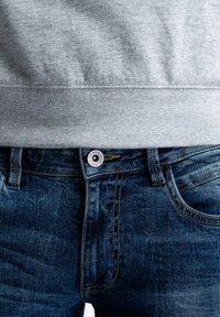 Manžety šedé mikiny nad tmavě modrými džínami s viditelným prošitím a zapínáním na knoflíky. Látka působí měkce a má mírnou strukturu.