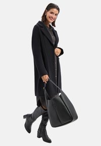 Zwarte wollen blend overjas met gestructureerde revers, knoopdetails en een losse pasvorm; gecombineerd met een bijpassende zwarte leren hobo tas en kniehoog laarzen.