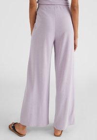 Pantalons larges en lavande clair, fabriqués à partir d'un tissu texturé. Taille élastique et ourlets lisses. Portés avec des sandales plates.