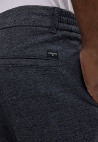 Graue Textilhose mit entspannter Taille, ausgestattet mit einer Gesäßtasche und einem kleinen Markenetikett. Subtiles Muster sorgt für visuelles Interesse.