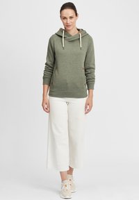 Felpa verde oliva con cappuccio, aperture laterali, dettagli con cordini e polsini a costine, abbinata a pantaloni bianchi a gamba larga e sneakers beige.