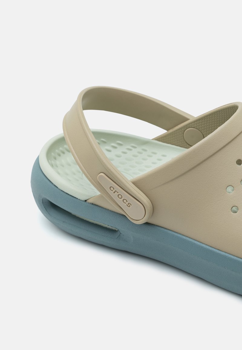 Beige Gummisandale mit blauer Sohle, ausgestattet mit einem Riemen und kleinen Belüftungsöffnungen. Texturiertes Innenleben und ein Logoakzent am Riemen.