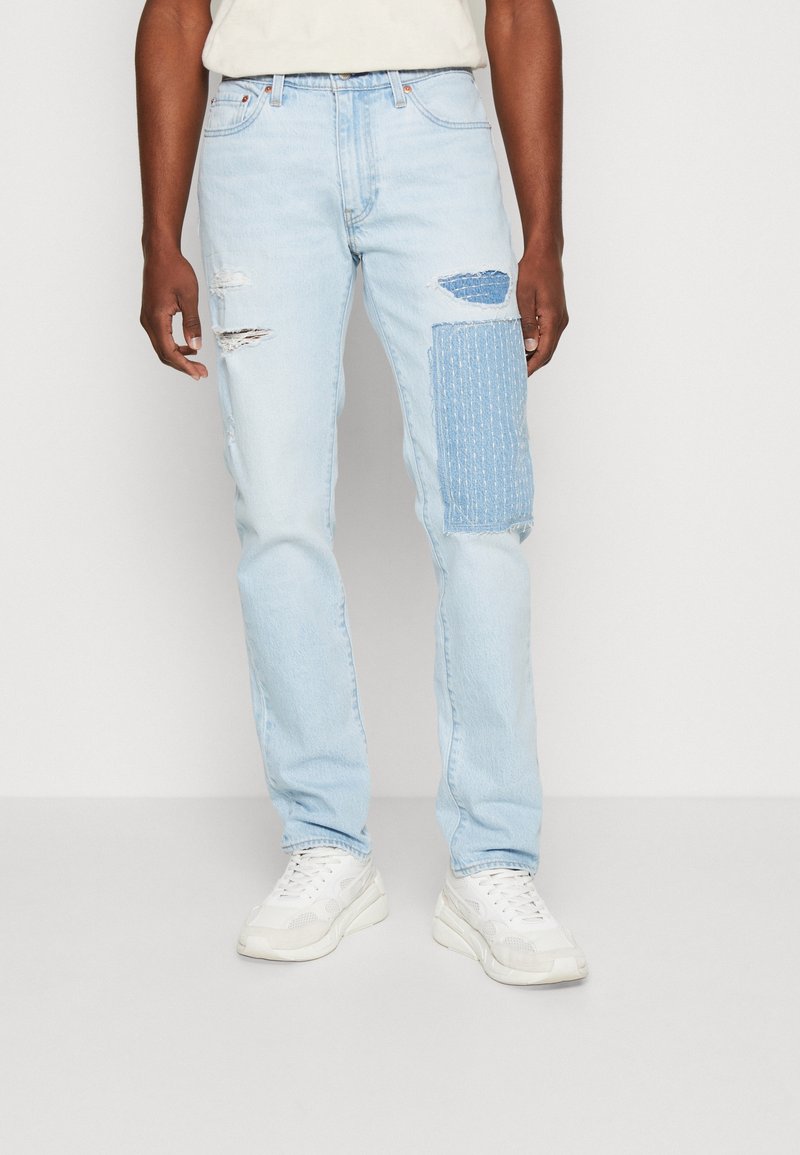 Ljusblå slitna jeans i denim, med rivningar och en strukturerad lappar på högra låret. Matchas med vita sneakers.