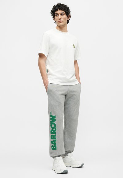 Νέος άνδρας με σγουρά μαλλιά, που φοράει λευκό t-shirt και γκρι φόρμα με πράσινο κείμενο «BARROW», στέκεται με τα χέρια στις τσέπες.