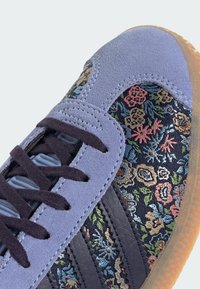 Purple suede sneaker with floral pattern textile, dark blue leather accents, and black laces. Translucent rubber outsole and detailed stitching.
