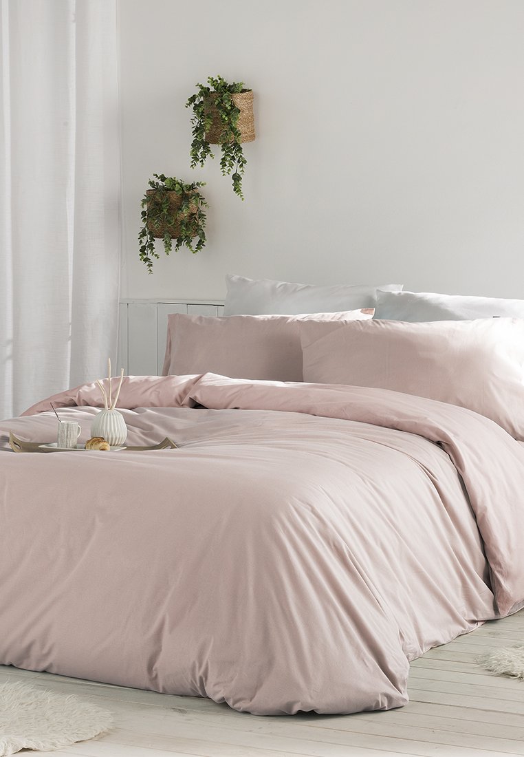 Leicht rosa Bettbezugset mit glatter Textur, begleitet von grauen Kissenbezügen; dekorative Pflanzen in geflochtenen Körben an der Wand.