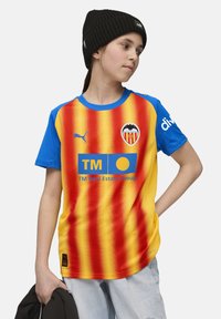 Ungdom som bär Valencia CF:s fotbollströja med röda och gula vertikala ränder, blå ärmar och en svart mössa, medan han håller en svart jacka.