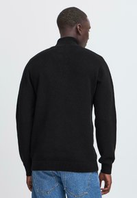 Svart strikkegenser med høy krage, lange ermer og ribbestrikkede mansjetter og nederkant. Brukt sammen med blå jeans, som viser en glatt tekstur.