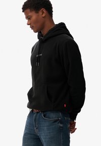 Sudadera de forro polar negra con un bolsillo frontal, capucha con cordón y detalle del logotipo. Se lleva con unos vaqueros de mezclilla azules, que tienen una etiqueta roja en el dobladillo.