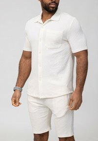 Chemise à manches courtes couleur crème avec un motif géométrique texturé et une poche poitrine, assortie à un short, le tout en tissu léger.