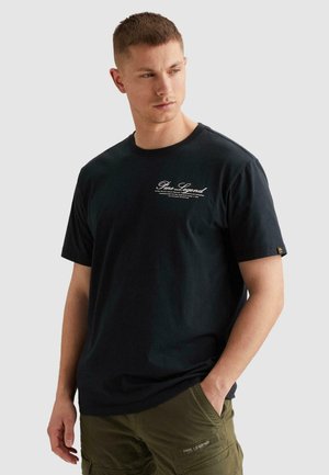 Muškarac u crnoj PME Legend majici i maslinasto zelenim cargo hlačama, gleda ulijevo s jednom rukom u džepu.