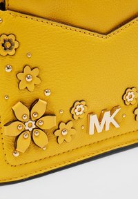 Bolso de mano de cuero amarillo con adornos florales, tachuelas doradas y un logotipo "MK" grabado en una superficie texturizada.