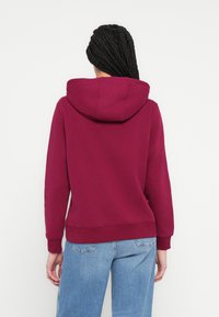 Personne avec des cheveux tressés portant un sweat à capuche rouge foncé et un jean bleu clair, de dos devant un fond blanc uni.