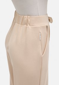 Pantalones beige con una cintura elástica, bolsillos laterales y una textura suave. Presentan una pequeña etiqueta cerca de la cintura.