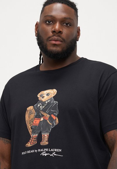 Melnas kokvilnas T-krekls ar grafiku, kurā attēlots ģērbts lācis ar saulesbrillēm, turot sniega kurpes, zem uzraksta "POLO BEAR by RALPH LAUREN."