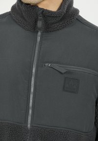 Musta fleece ja kangaspulloveri korkealla kauluksella, vetoketjullinen rintatasku ja kohopainettu logo. Teksturoitu fleece kauluksessa ja helmassa.