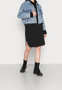 Denimová bunda s ošoupanými okraji, nošená přes černé šaty. Model má na sobě černé šněrovací boty se žlutou visačkou, což ukazuje na kontrastní textury.