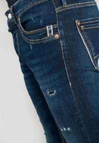 Jeans de mezclilla de color azul oscuro con un aspecto desgastado que presenta pequeños desgarros, costuras en contraste y un sutil detalle a rayas en los bolsillos.