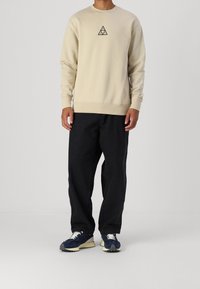 Felpa di cotone beige con un logo triangolare nero al centro, abbinata a pantaloni neri larghi e sneaker blu scuro.