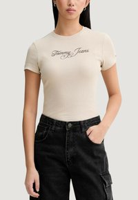 Béžové žebrované tričko s krátkými rukávy a logem "Tommy Jeans" psaným kurzívou. Kombinováno s černými džíny s pohodlným střihem.