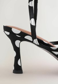 Sandale noire à talon haut avec des pois blancs, des fines brides et un talon effilé sur un fond neutre.