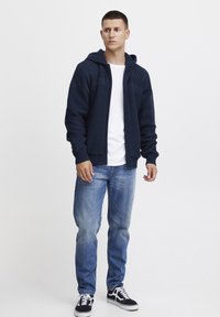 Jūrinės mėlynos spalvos megztas gobtuvu kardiganas su raukinėmis apykaklėmis, užtrauktuku ir tekstūrinės detalės, dėvimas ant baltos marškinėlės ir mėlynų džinsų.