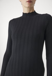 Top black cu mânecă lungă, cu textură ribbed, guler înalt și margini zimțate. Design ajustat care evidențiază silueta.