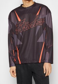 Långärmad svart sporttröja med orange detaljer och en logotyp. Har ett geometriskt mönster och en rund halsringning. Mjukt och andningsbart material.