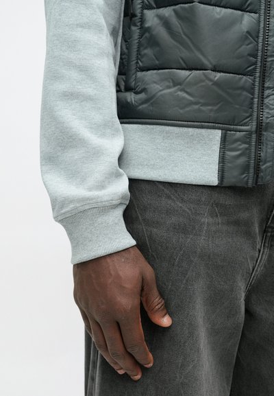 Pilkas vilnonis ilgomis rankovėmis marškinėliai su banguotais rankogaliais, derinami su juodu paminkštintu liemeniu. Tamsiai pilkos džinsai. Matoma ranka su tekstūruota oda.