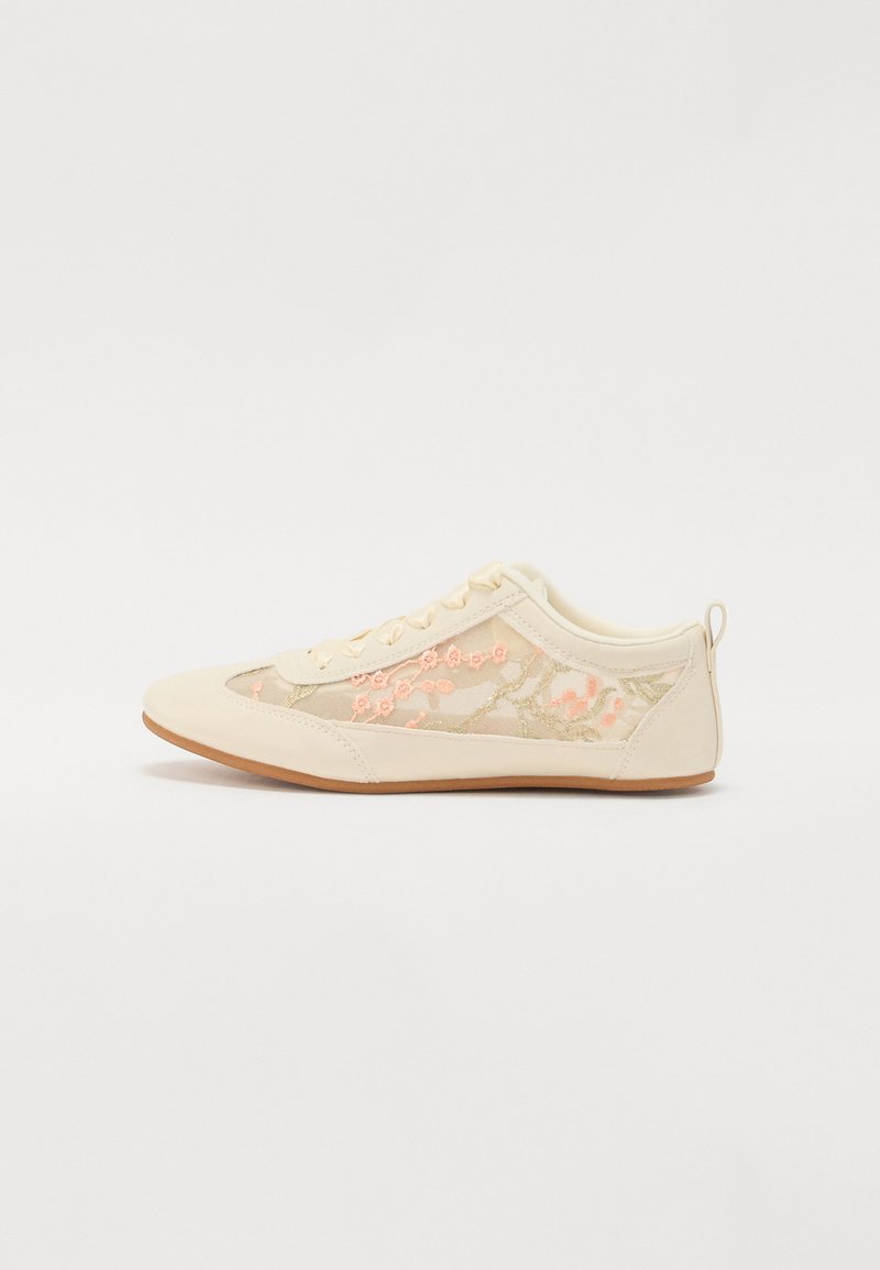 Sneaker color crema con pannelli laterali in rete ricamati a fiori, dettagli beige e suola in gomma marrone chiaro. Presenta una chiusura con lacci e un anello posteriore.