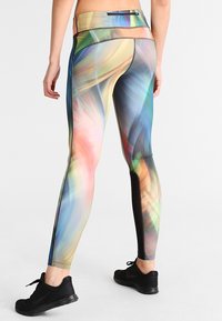 Osoba nosząca legginsy w wielokolorowy, abstrakcyjny wzór oraz czarne buty sportowe, stojąca plecami do kamery na białym tle.